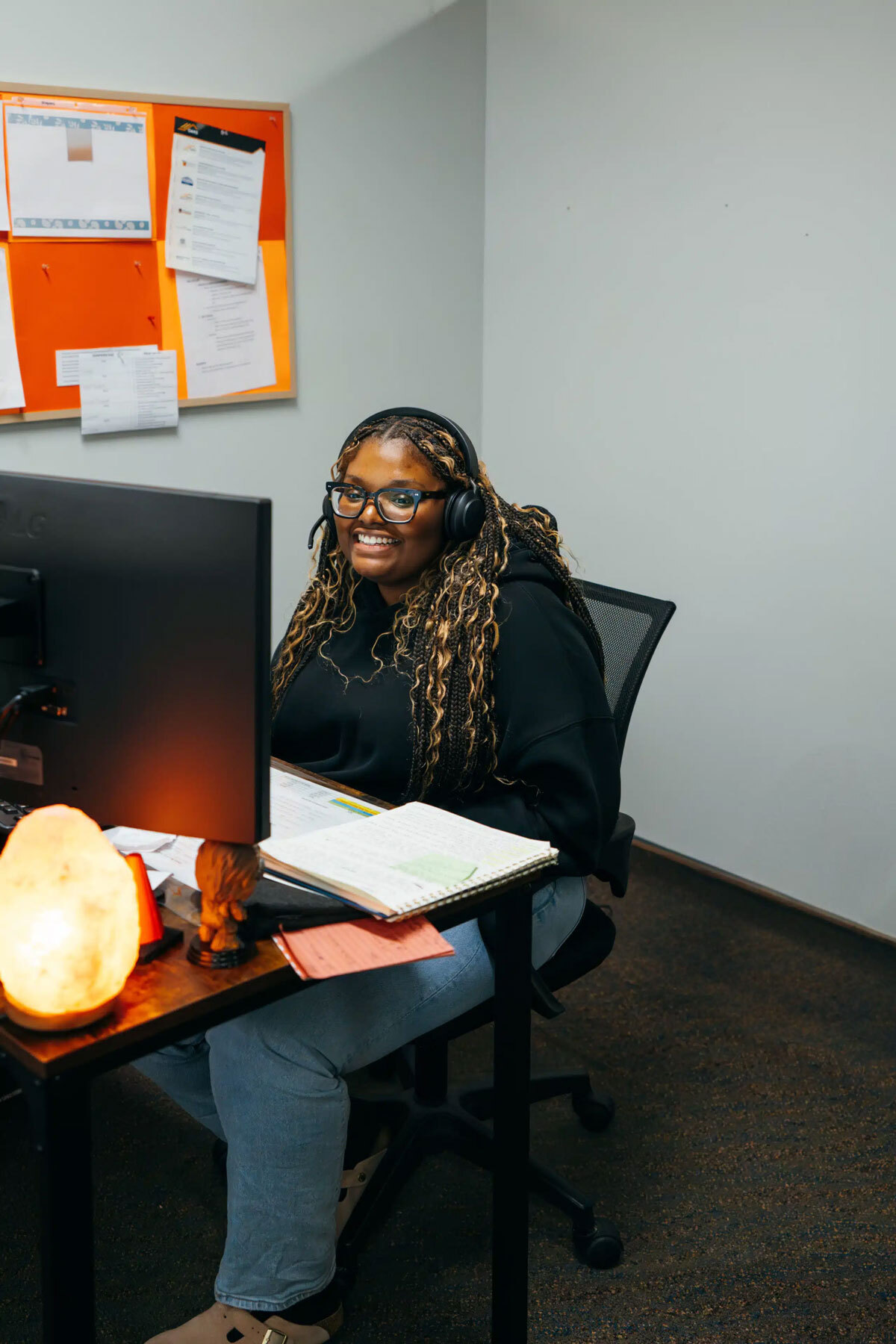 A woman in a black headset sits at a desk with a computer, notebook, and lamp. She appears to be in an office setting.