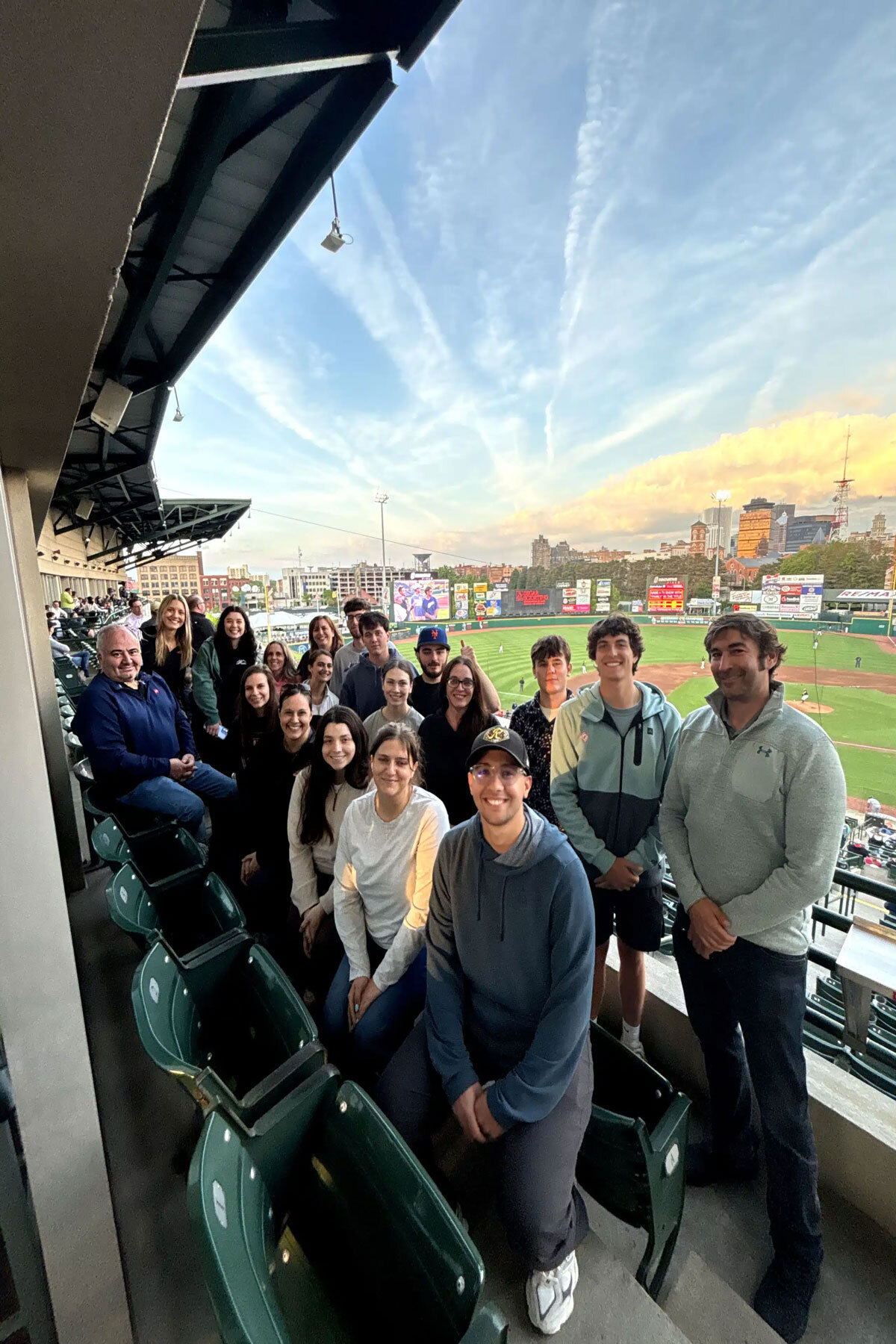 A group of people smiling and posing for a photo at a sports stadium. They appear to be in a suite or VIP area.