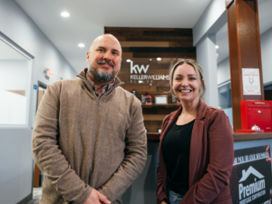 A man and woman standing side by side in a real estate office. They are smiling and looking at the camera.