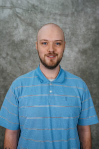 A man with a shaved head and short beard wearing a blue polo shirt. He is standing in front of a gray background.