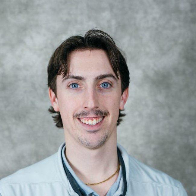 Portrait of a smiling man with a mustache. He is wearing a gold necklace.