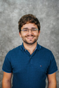 A young man with short brown hair and glasses smiling at the camera. He is wearing a blue polo shirt.
