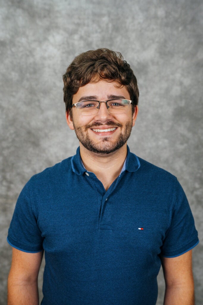 A young man with short brown hair and glasses smiling at the camera. He is wearing a blue polo shirt.