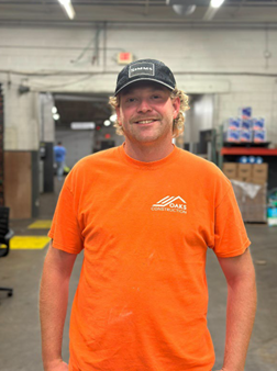 Image shows a man wearing a bright orange shirt and a black cap, standing in a warehouse with items in the background.