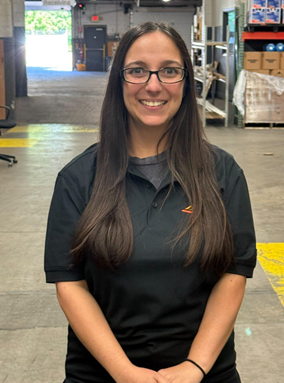 A smiling woman with glasses and long brown hair stands in a warehouse. She is wearing a black polo shirt with an orange logo.