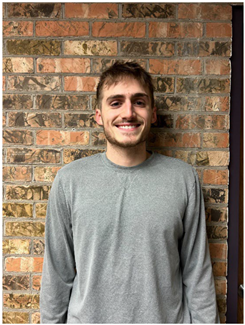 A young man with light brown hair and beard, wearing a gray sweatshirt, stands smiling against a rustic brick wall. The natural lighting accentuates his cheerful expression.