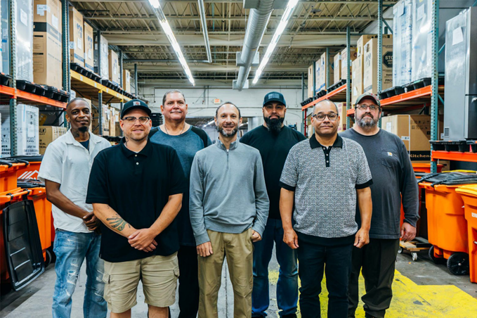 A group of warehouse employees standing together surrounded by tall shelving units filled with boxes and orange containers.