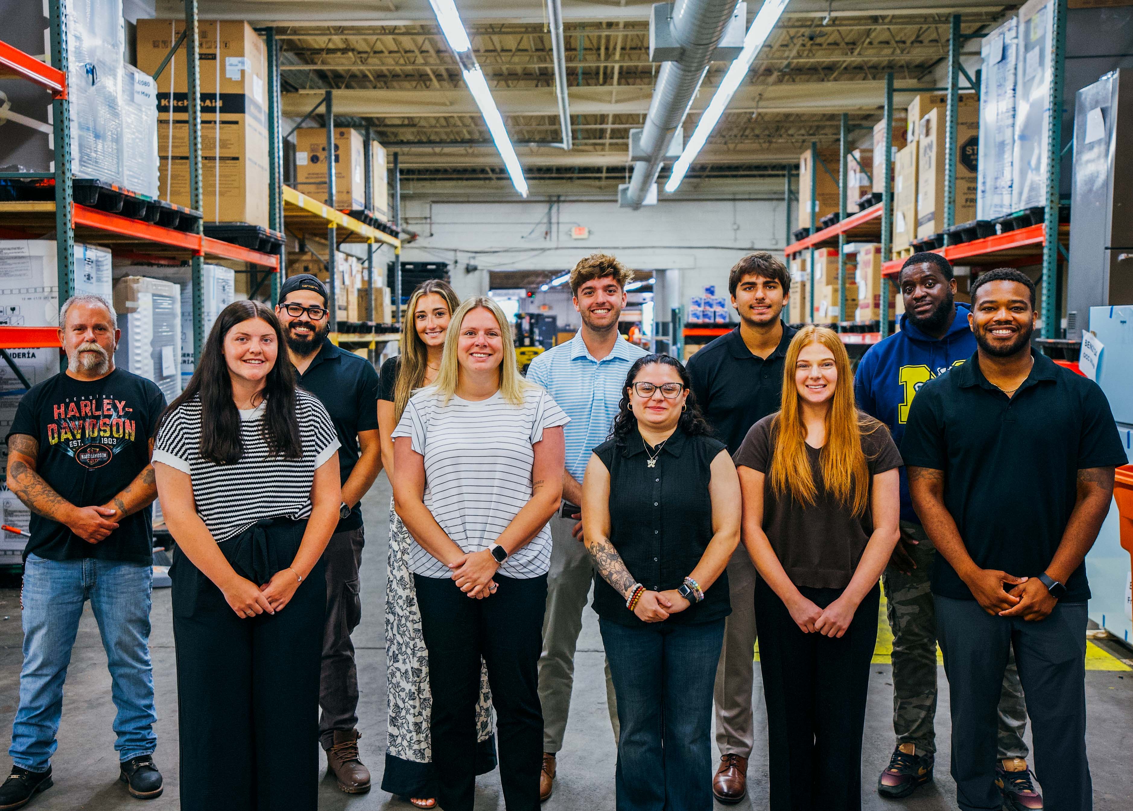 A group of people standing in a warehouse filled with boxes. The team is smiling and appears to be working together.