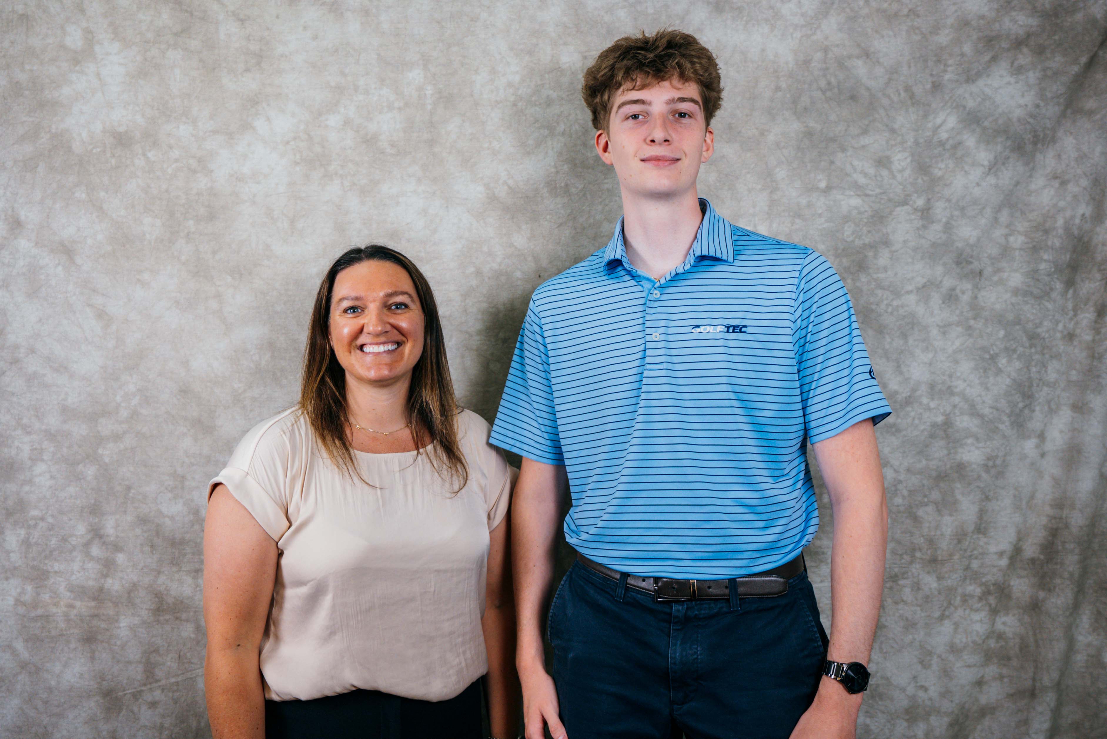 A woman in a cream top stands beside a young man in a blue striped shirt against a textured backdrop.