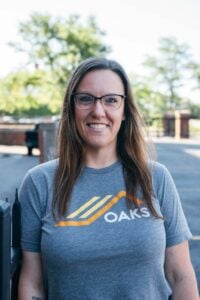 A smiling woman with glasses wearing a gray T-shirt with the 'OAKS' logo.