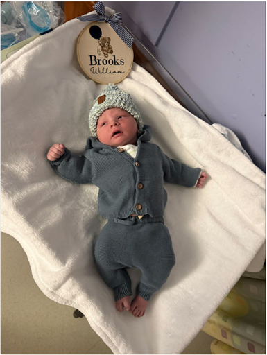 A newborn baby named Brooks William lies on a white blanket wearing a blue onesie and a knit hat with a bear design, surrounded by baby gifts.