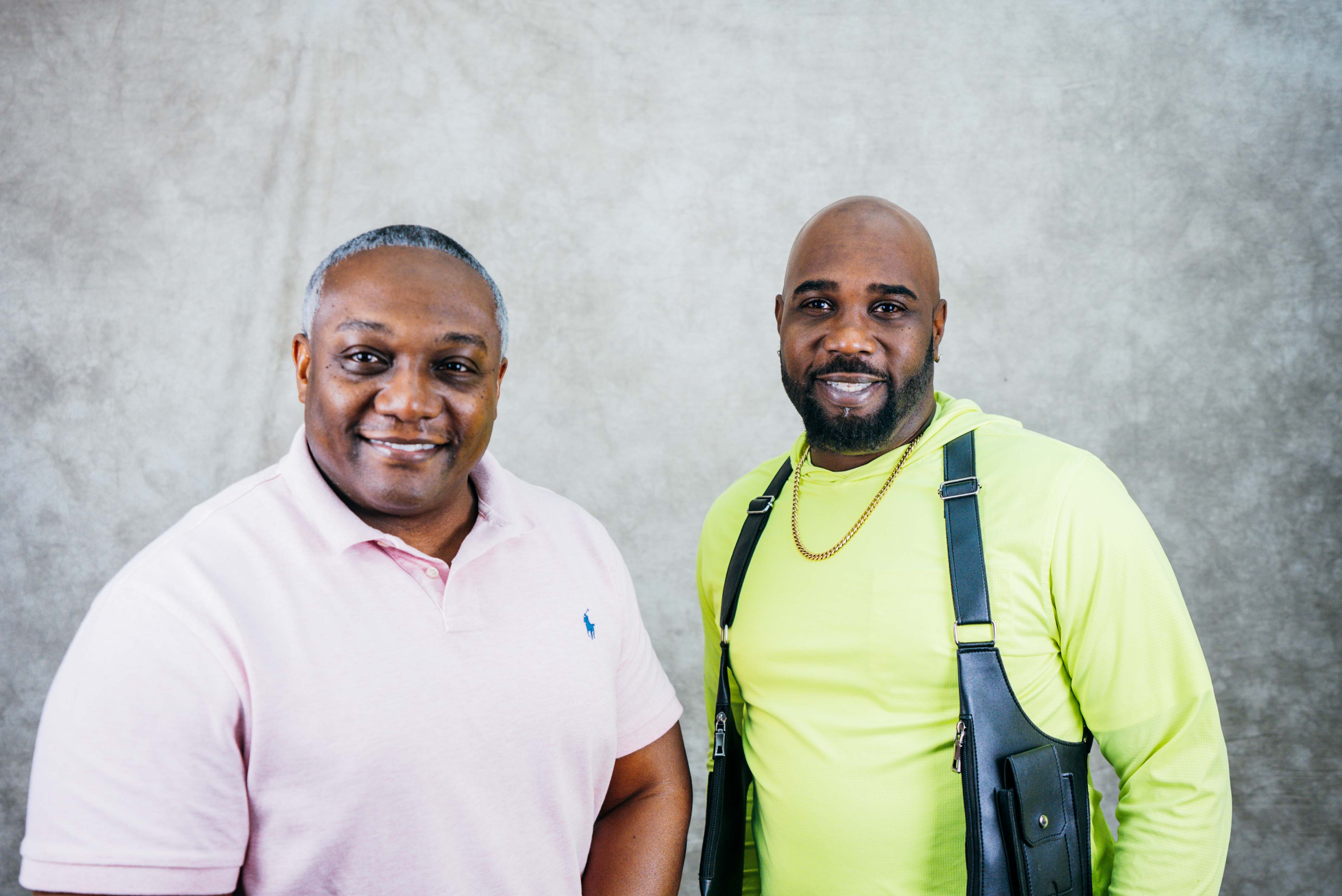 Two casually dressed men smiling at the camera. One wears a pink polo and the other a neon yellow shirt with a crossbody bag.