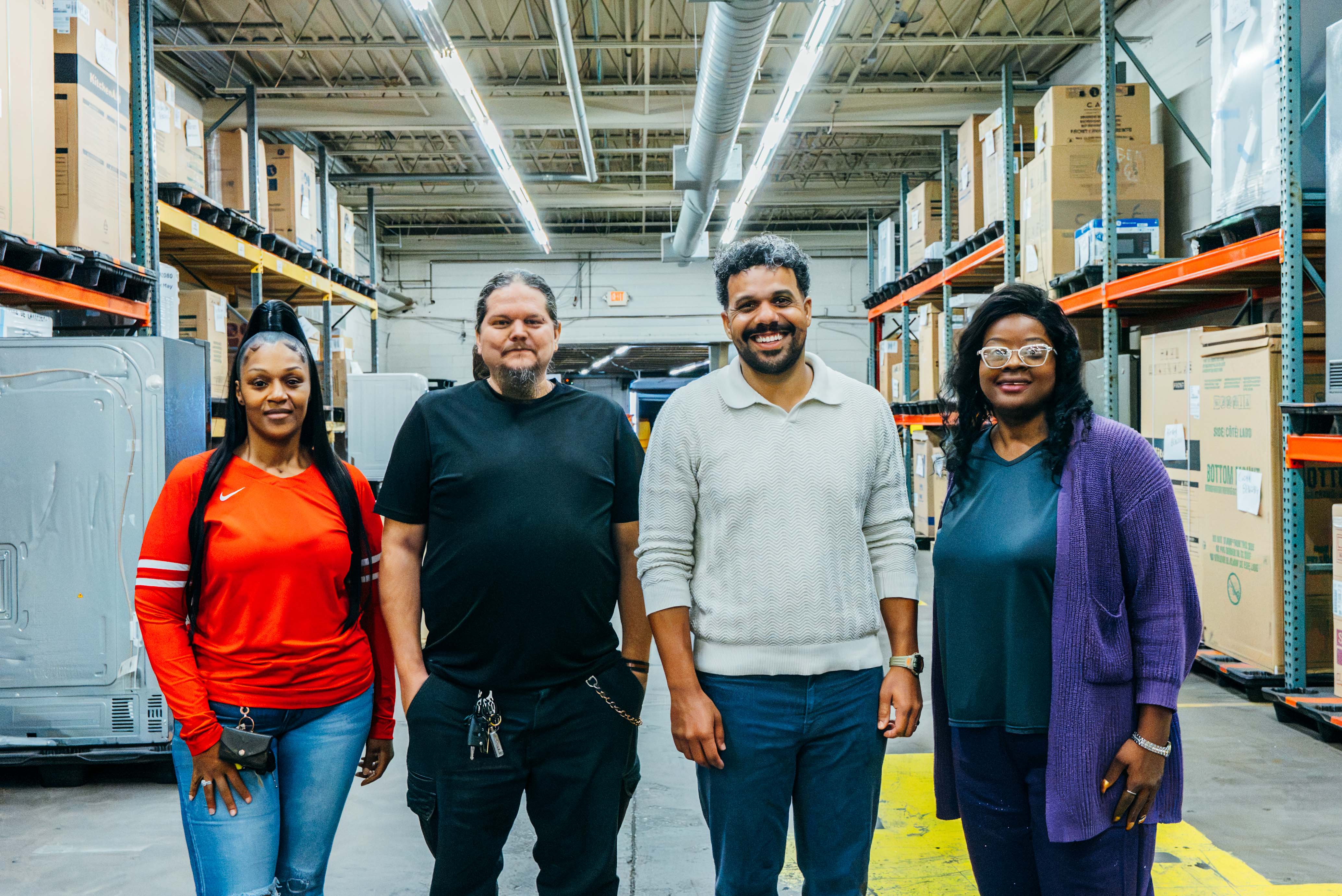 A diverse team of four warehouse workers stands side by side in a well-lit industrial facility, surrounded by tall storage racks and boxes.