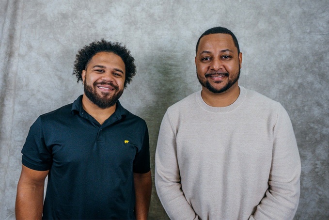 Two smiling men standing together against a grey textured wall, featuring dark and light clothing in complementary tones.