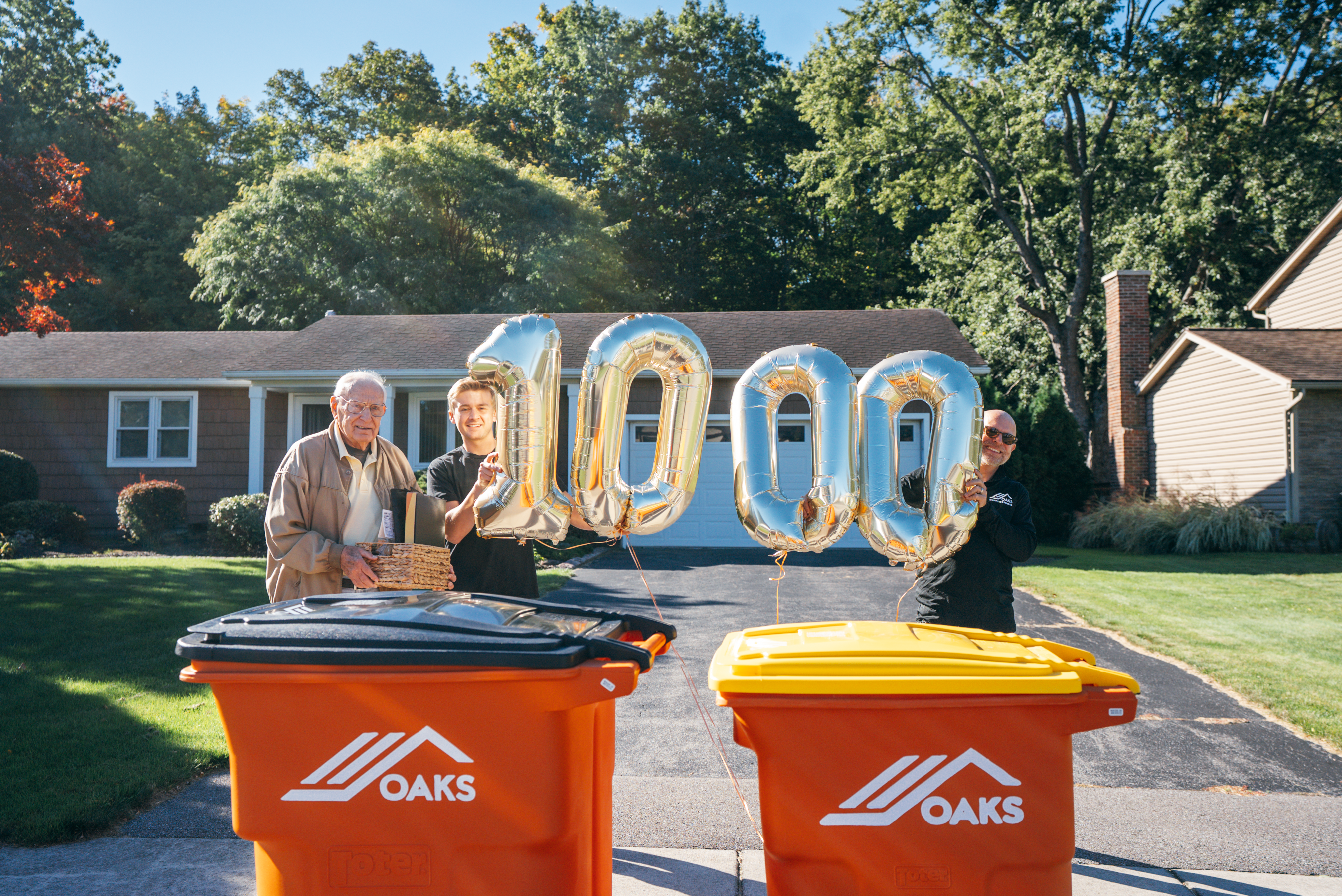 Family celebrating with a 100th anniversary balloon in front of two orange Oaks trash bins in a suburban driveway.