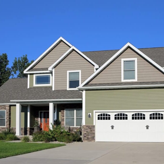 A two-story house with green siding and a white garage door. The house has a covered porch.