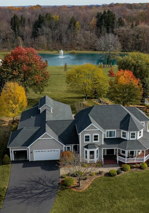 A large house with a gray roof and white walls sits on a green lawn. The house has a two-car garage.