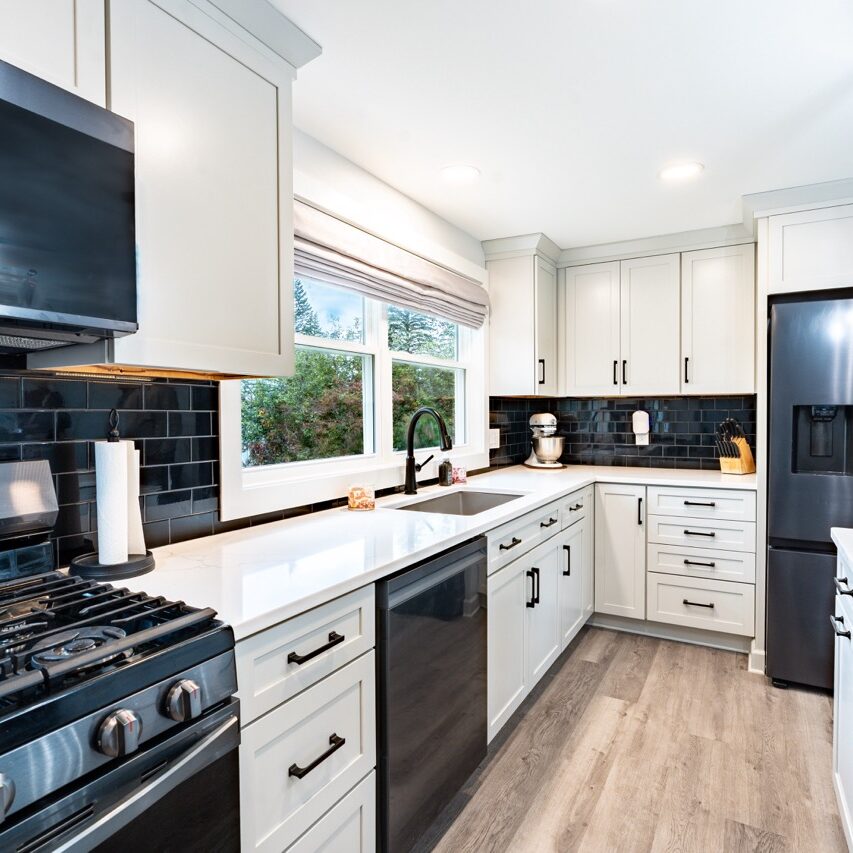 A modern kitchen with white cabinets and black appliances. The kitchen has a large window above the sink.